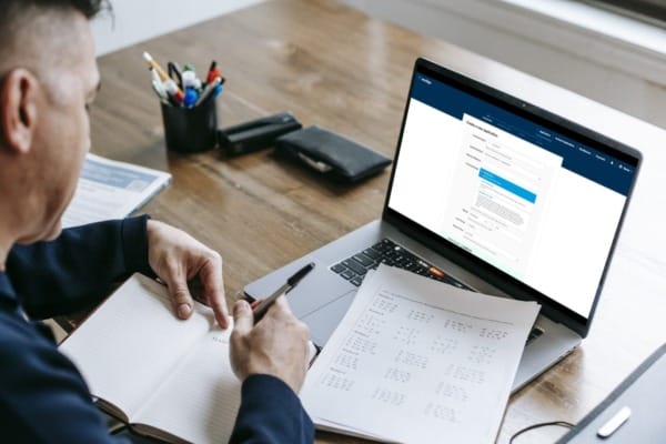 A man sits at a wooden desk, taking notes in a notebook while reviewing financial information on a laptop screen. The laptop displays an online finance application form, indicating a kitchen loan or home improvement financing process. Nearby, a document with calculations is visible, suggesting careful planning of loan terms or repayment options. The setting conveys a focus on managing kitchen loan finances and making informed financial decisions.