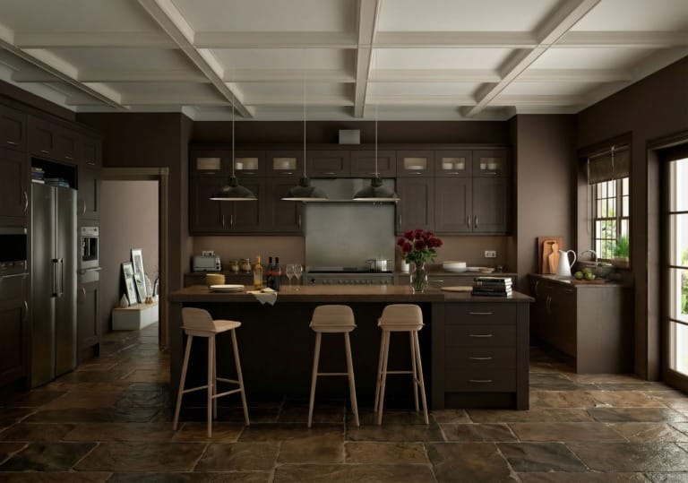 A warm, modern kitchen with dark wood cabinets, a large central island, and three light wooden bar stools. Three industrial pendant lights hang above the island, which is topped with wine bottles, glasses, plates, books, and a vase of red flowers. Stainless steel appliances line the back wall, and a window on the right lets in natural light with greenery visible outside. The floor is made of large rustic stone tiles, adding texture to the space.