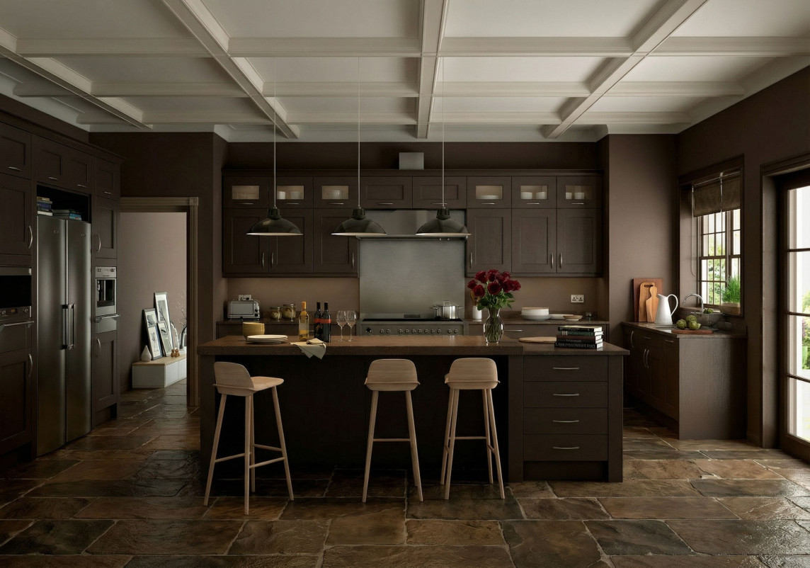 A warm, modern kitchen with dark wood cabinets, a large central island, and three light wooden bar stools. Three industrial pendant lights hang above the island, which is topped with wine bottles, glasses, plates, books, and a vase of red flowers. Stainless steel appliances line the back wall, and a window on the right lets in natural light with greenery visible outside. The floor is made of large rustic stone tiles, adding texture to the space.