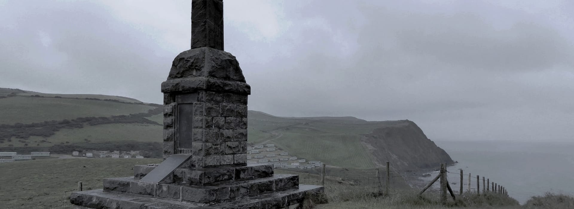 weathered stone monument on grassy cliff overlooking grey sea and cloudy sky