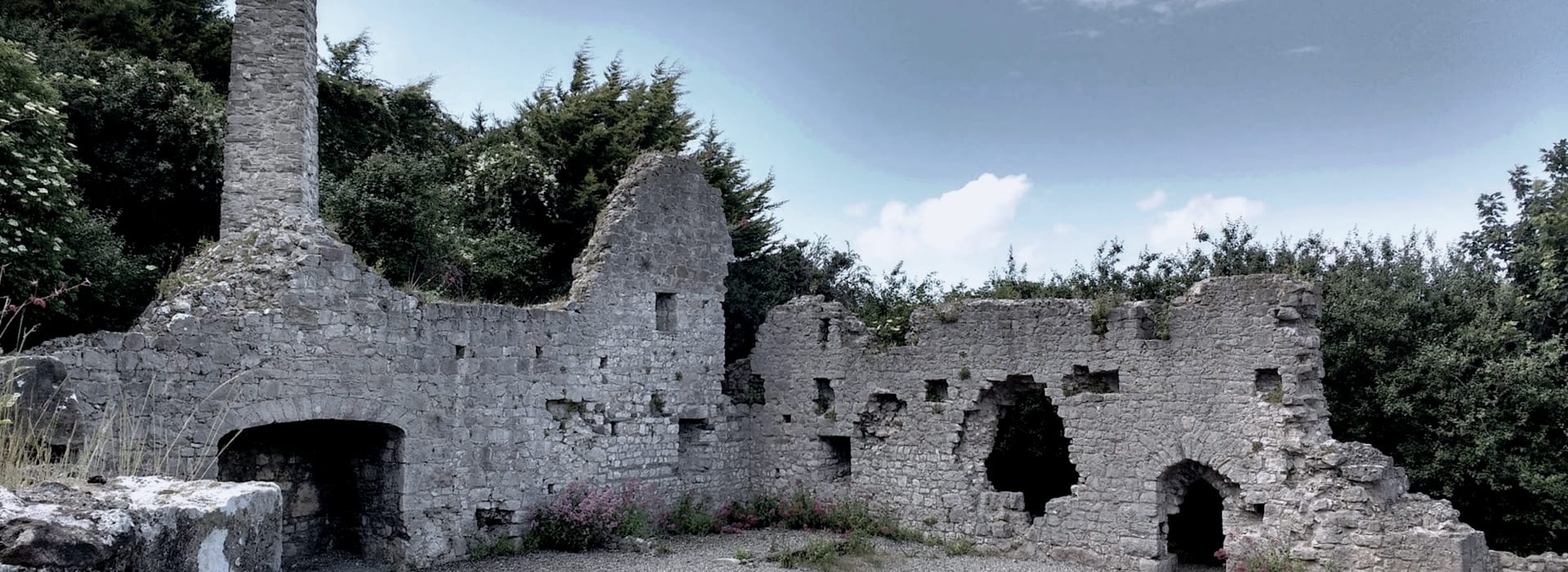 weathered stone ruins with arched openings and chimney against green trees