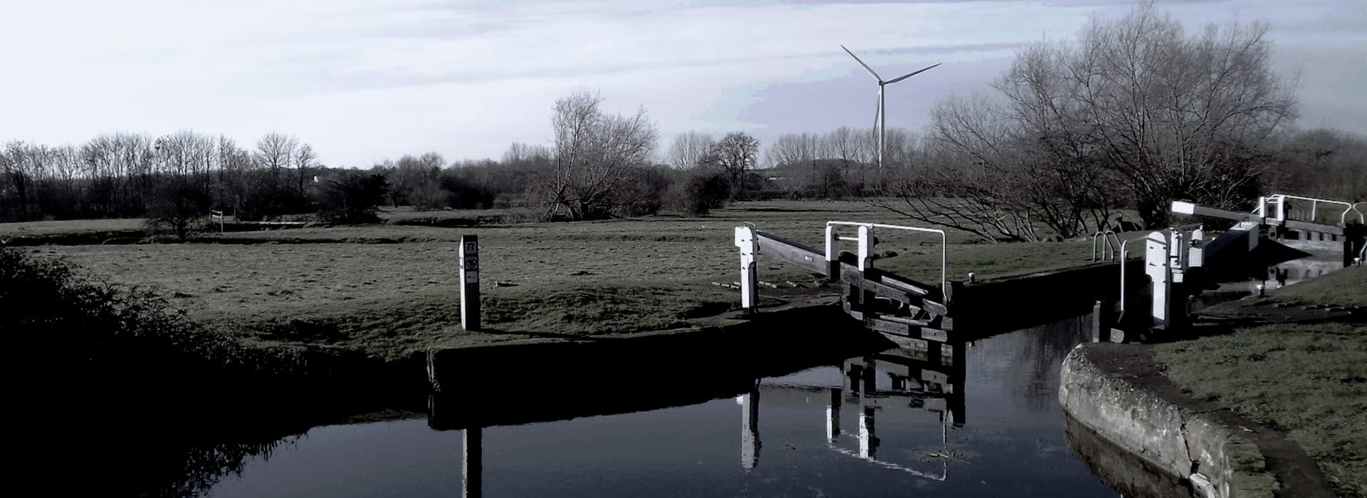 rural canal lock with white gates and distant wind turbine under grey sky