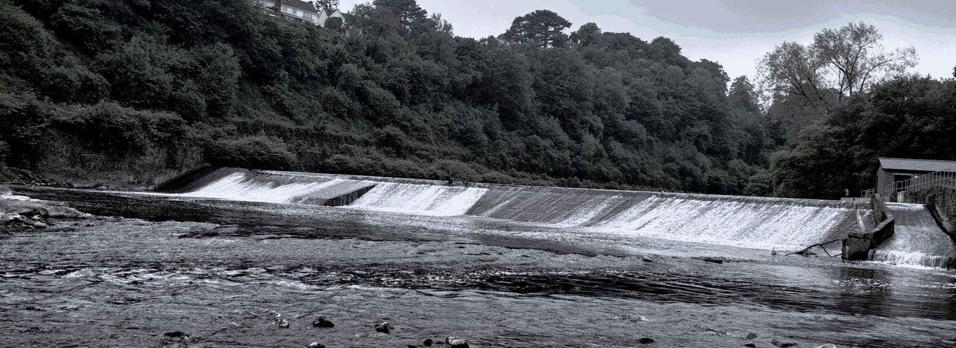 wide concrete weir with cascading water, surrounded by dense green woodland