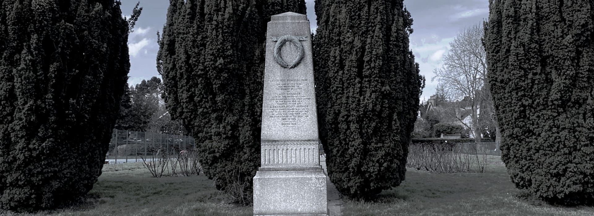 grey stone memorial obelisk with wreath carving, flanked by tall dark trees