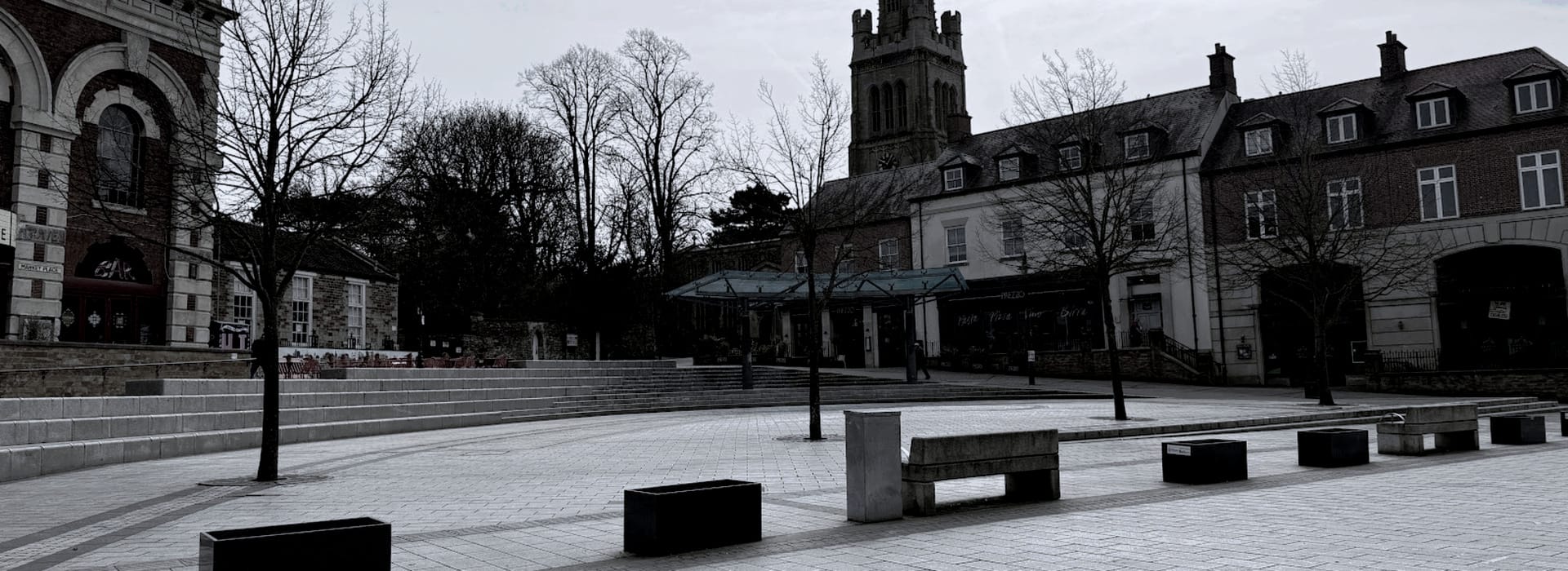 Grey paved town square with bare trees and stone benches, church tower in background