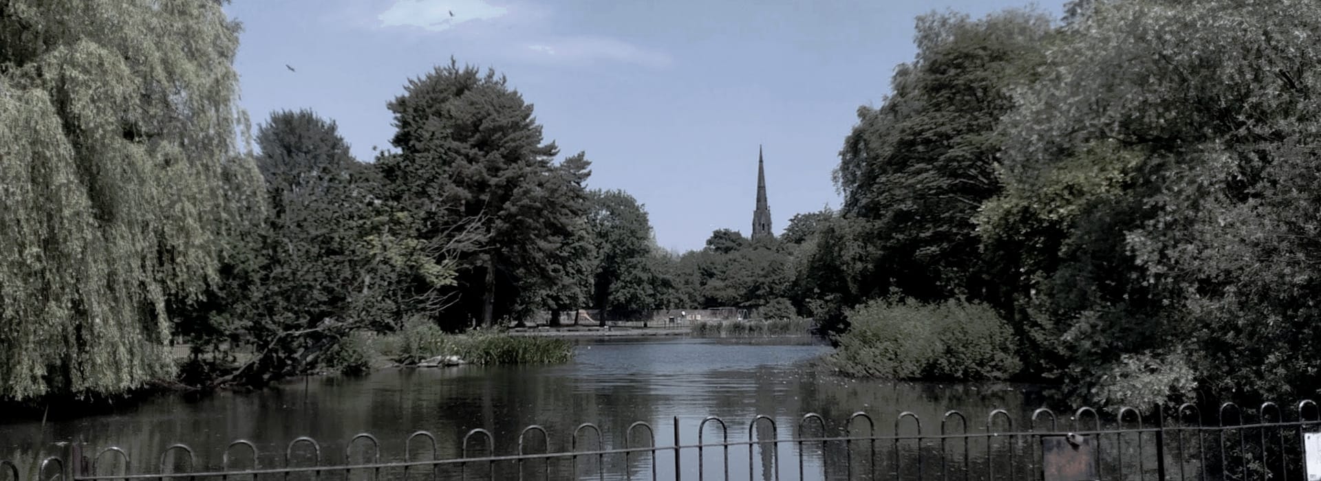 lake with overhanging trees and distant church spire under pale blue sky