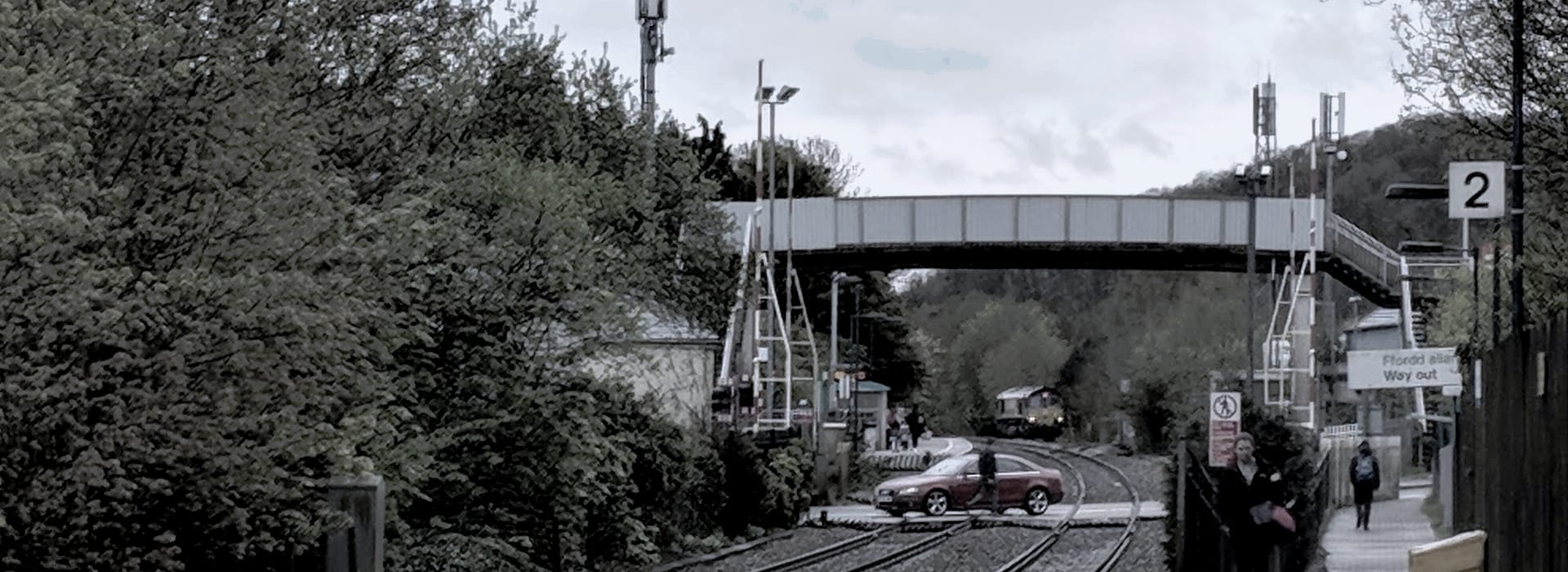 Muted railway scene with grey footbridge, red car at level crossing, overcast sky