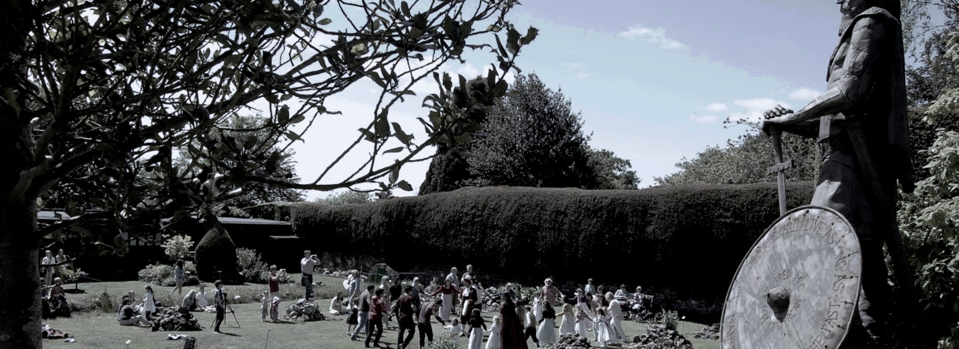 large metal statue with shield overlooking garden with people and manicured hedges