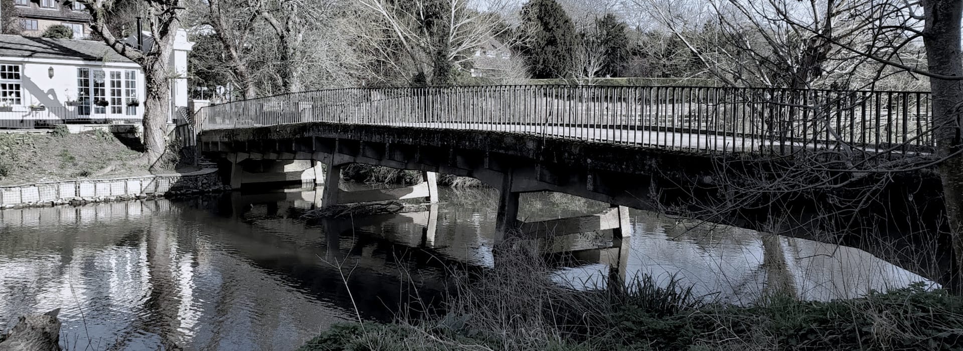 concrete footbridge with metal railings over calm river, winter trees surrounding