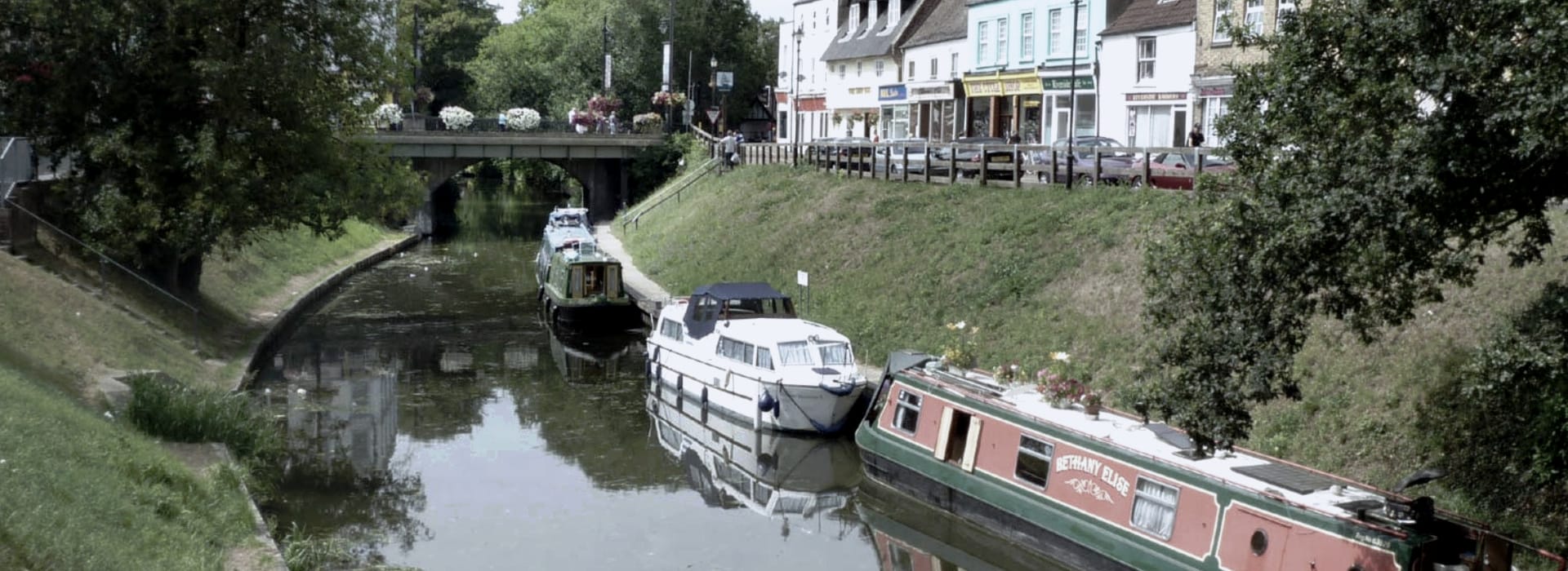 moored canal boats beside grassy bank and colourful shopfronts under leafy trees