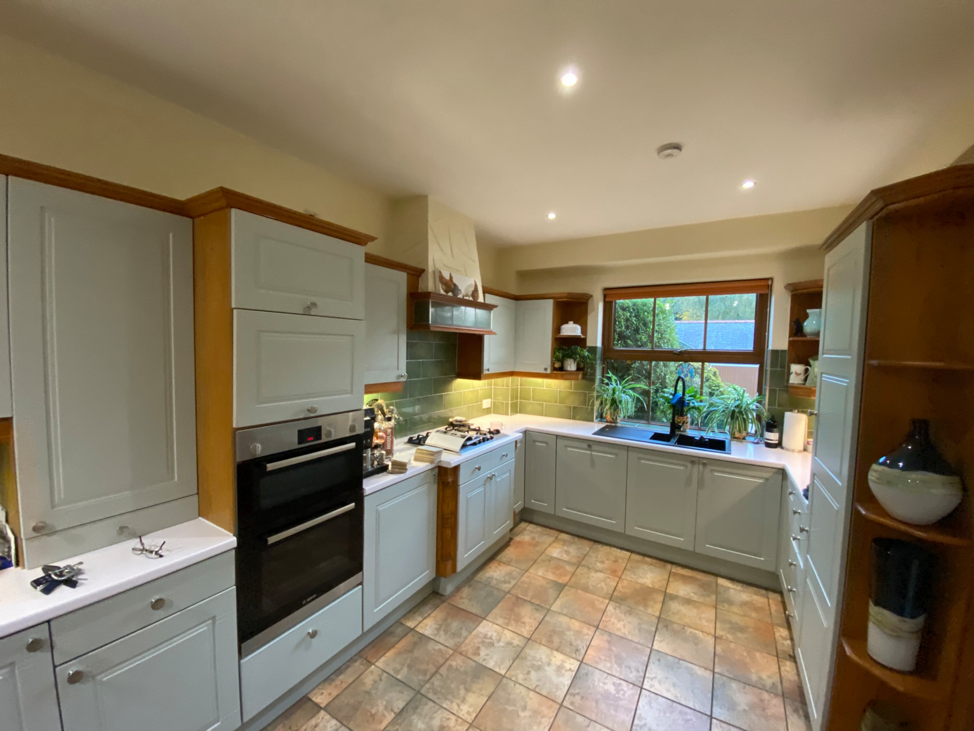 Maree Before L-shaped kitchen with pale green shaker cabinets, contrasting wood trim, white worktops, and green splashback tiles