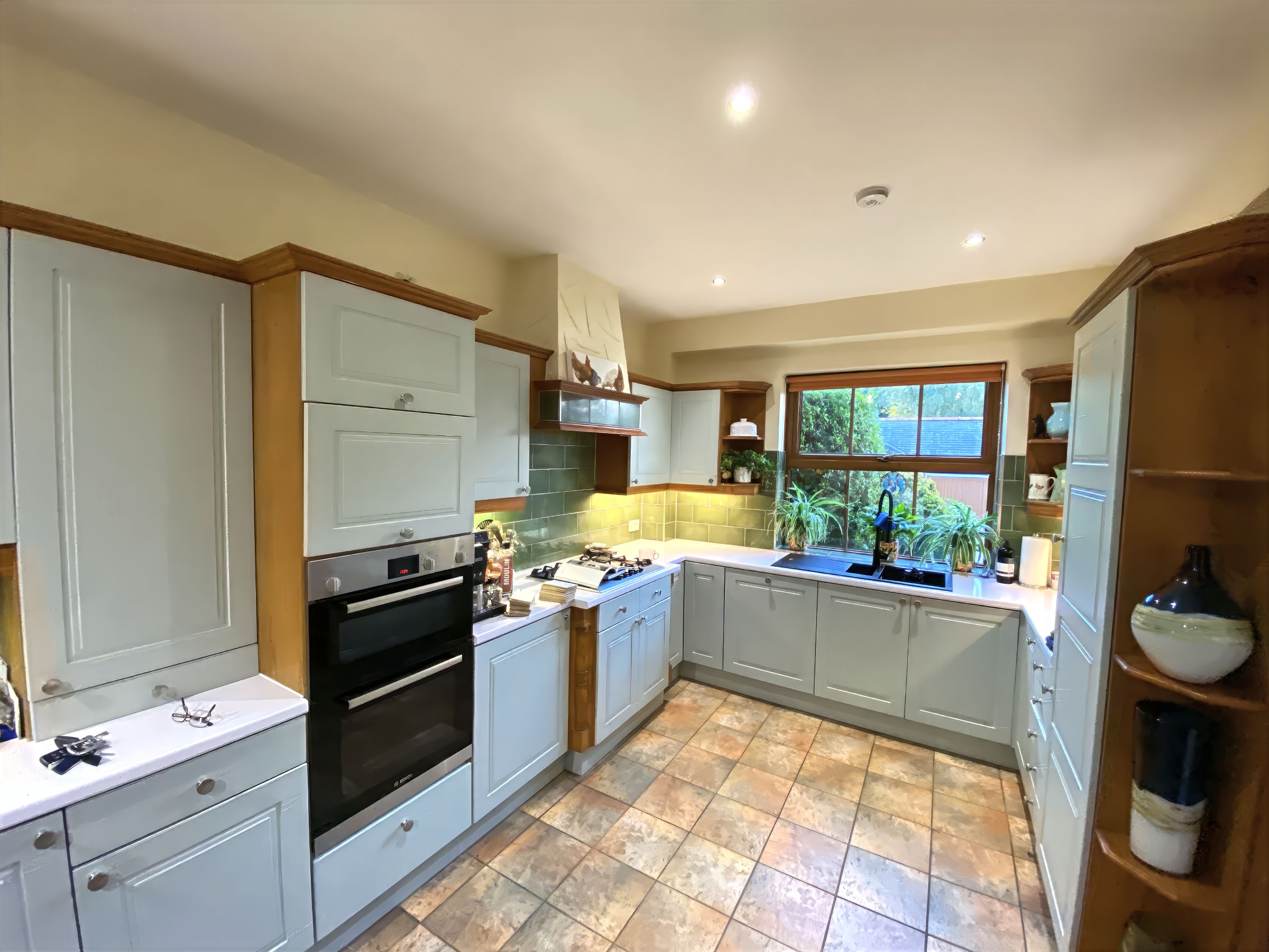 L-shaped kitchen with pale blue shaker cabinets, wood trim, green tiled splashback and terracotta-style floor tiles